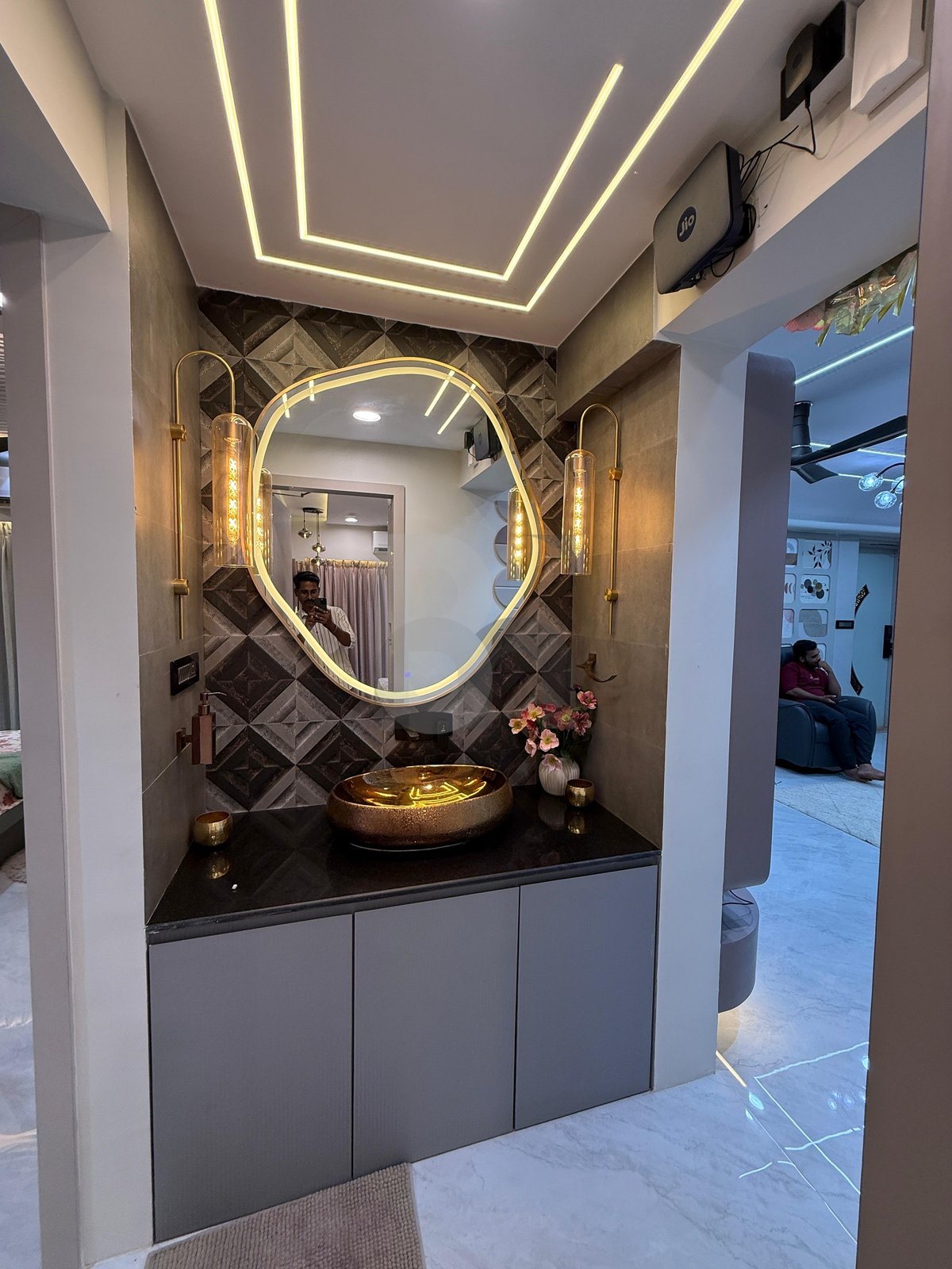 Dining foyer hand wash area with asymmetric illuminated mirror, black granite counter, and geometric cove ceiling by Brand Meter